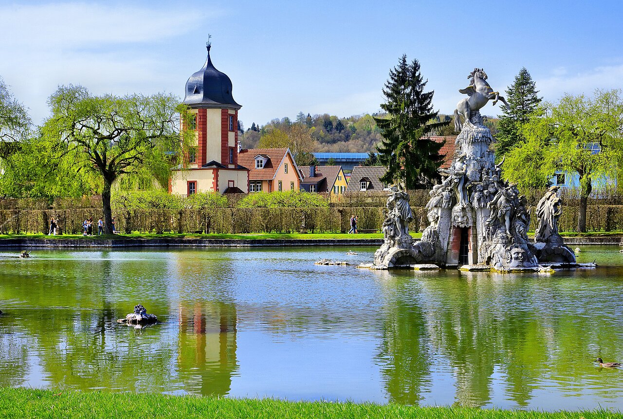 Aus dem großen See im Hofgarten erhebt sich die Skulptur des Musenbergs Parnass, im Hintergrund der Wasserturm