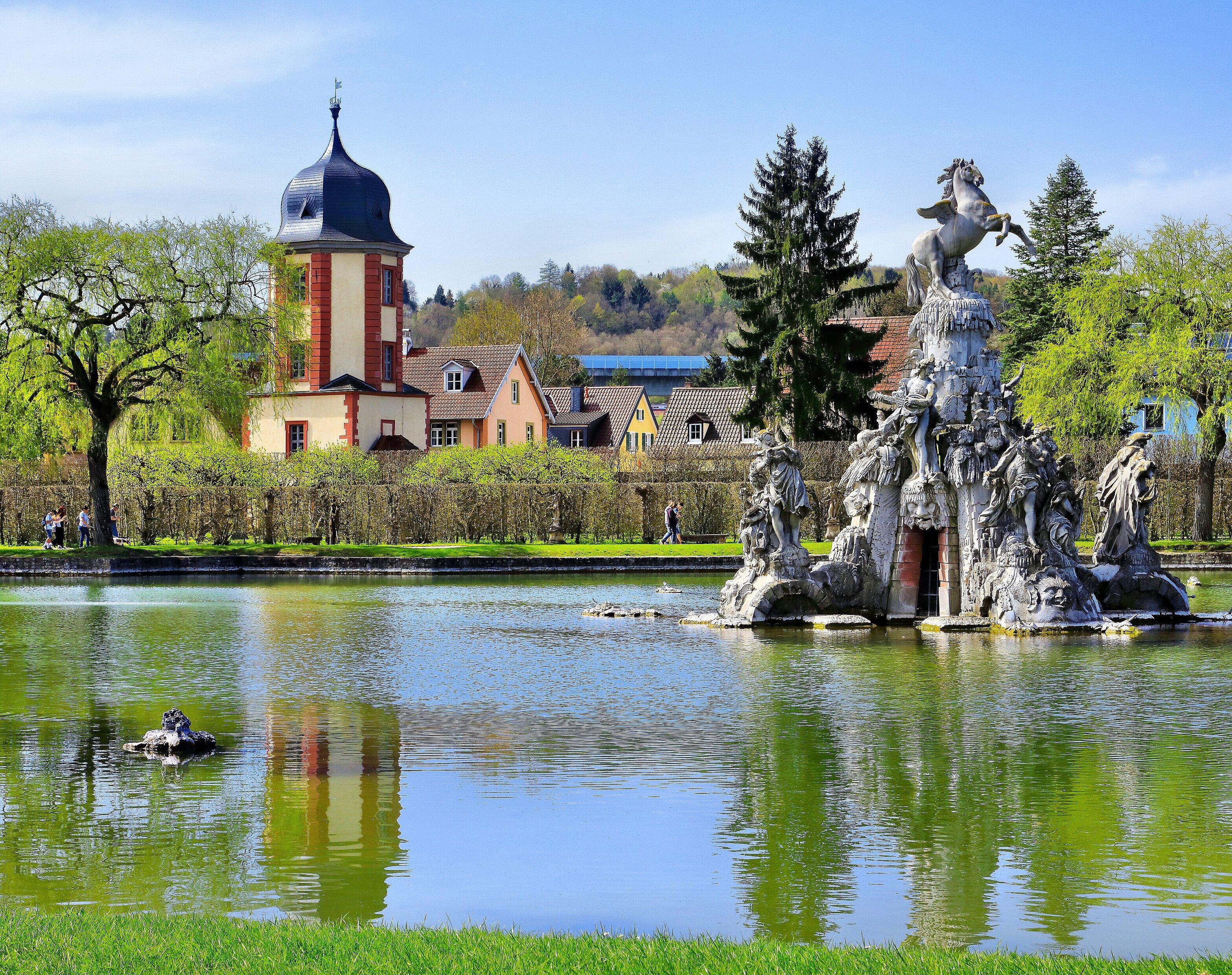 Aus dem großen See im Hofgarten erhebt sich die Skulptur des Musenbergs Parnass, im Hintergrund der Wasserturm