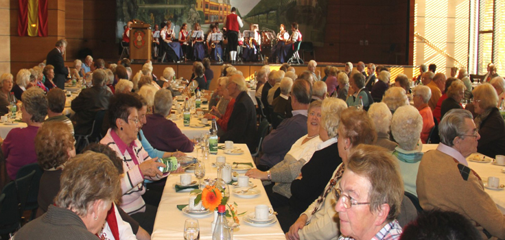 An den Tischen in Saal sitzen zahlreiche Seniorinnen und Senioren bei einem Glas Wein oder Wasser. Im Hintergrund spielt der Musikverein auf der Bühne.